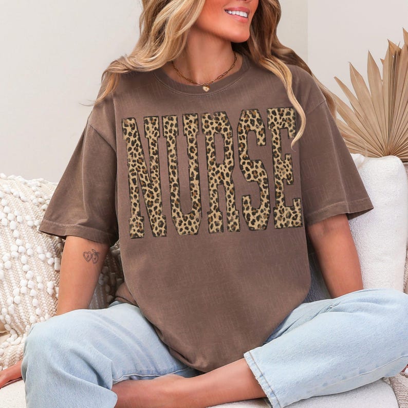 May include: A brown t-shirt with the word "NURSE" in large leopard print letters. The shirt has short sleeves and a relaxed fit. The person in the image is wearing light blue jeans.