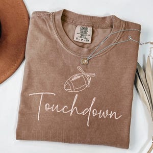 May include: A brown Comfort Colors t-shirt with a white football graphic and the word "Touchdown" in a script font. A silver necklace with a pendant is draped across the shirt.
