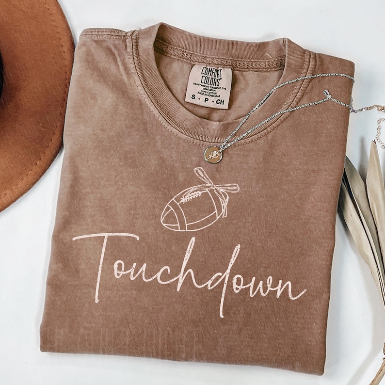 May include: A brown Comfort Colors t-shirt with a white football graphic and the word "Touchdown" in a script font. A silver necklace with a pendant is draped across the shirt.