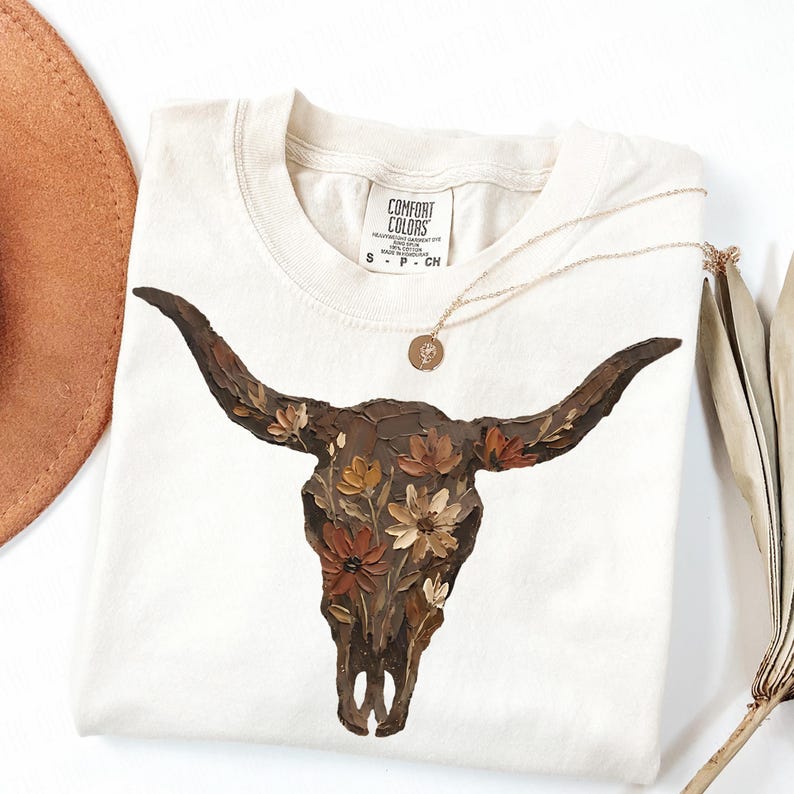 May include: Cream-colored t-shirt featuring a detailed illustration of a bull skull adorned with flowers in shades of brown, orange, and cream. A gold necklace with a pendant rests on the shirt. The shirt has the "Comfort Colors" brand label.