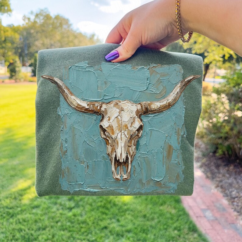 May include: A square, olive green fabric with a textured, teal blue painted square featuring a detailed, metallic gold-coloured bull skull. The skull has long horns and is the focal point of the artwork. The piece is held by a hand with purple nail polish.