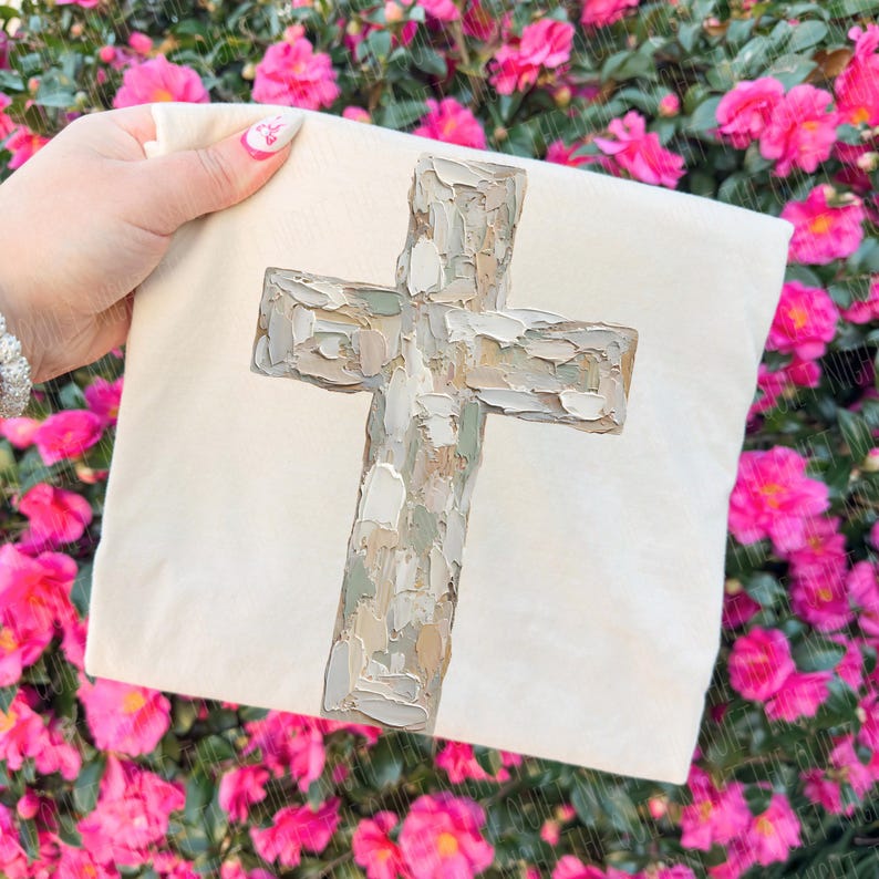 May include: A cream-colored square pillow featuring a textured, painted cross in shades of gray, beige, and green. The cross is the focal point, set against a plain background. The pillow is held in front of a backdrop of pink flowers.