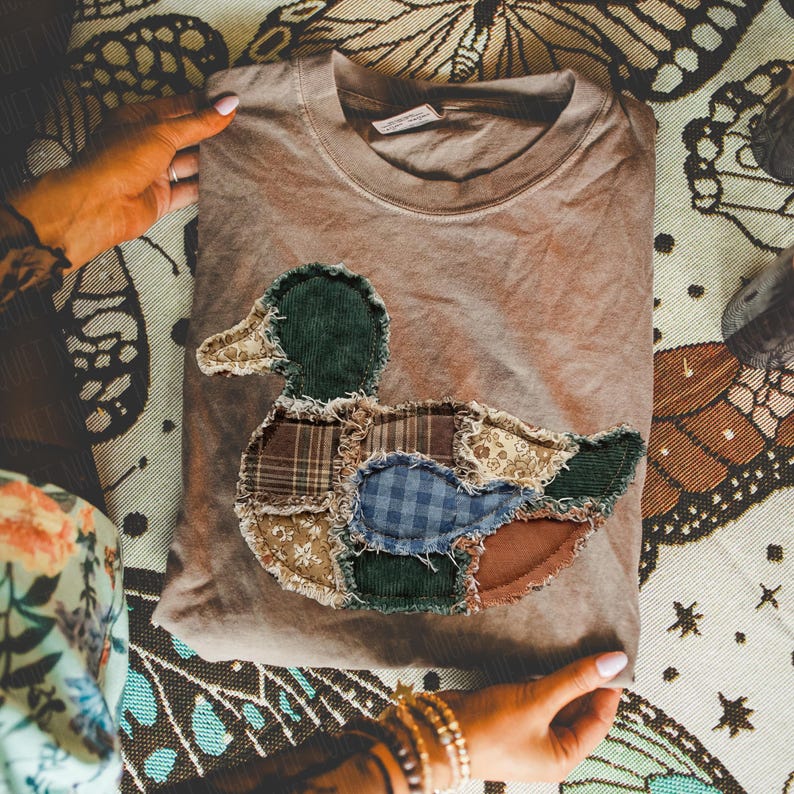 May include: A light brown crew neck t-shirt featuring a large duck-shaped appliqu&eacute; on the front. The duck is constructed from various fabric patches including dark green corduroy, blue gingham, brown plaid, and floral patterns with frayed edges.