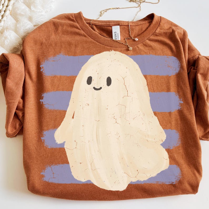 May include: A rust-colored t-shirt featuring a central graphic of a cream-colored ghost with a simple smiling face. The ghost is layered over four horizontal, distressed light purple brushstroke stripes. The shirt has a crew neckline.