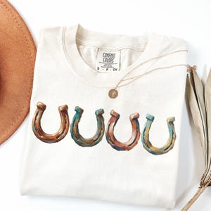 May include: Cream-colored t-shirt featuring four colorful horseshoe graphics. The horseshoes are painted in shades of brown, blue, and orange. A gold necklace with a pendant rests on the shirt. The shirt has a Comfort Colors tag.