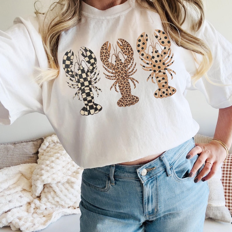 May include: White t-shirt featuring three lobster designs. The lobsters have different patterns: black and white checkered, leopard print, and brown with black spots. The shirt is worn with blue jeans.