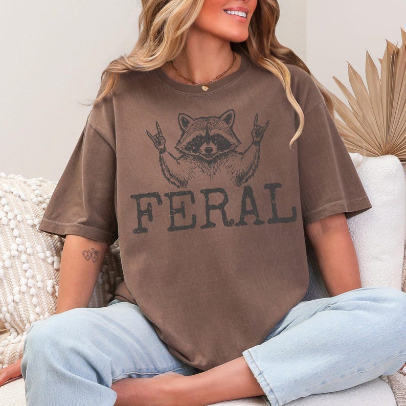 May include: Brown t-shirt featuring a raccoon graphic with the word "FERAL" in a distressed font. The raccoon is depicted with its hands in a rock-and-roll gesture. The shirt has short sleeves and a relaxed fit.