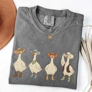 May include: A gray Comfort Colors t-shirt featuring four cartoon geese wearing cowboy hats and boots. The geese are in various poses, with different colored hats and boots. A silver necklace with a small pendant is on the shirt.