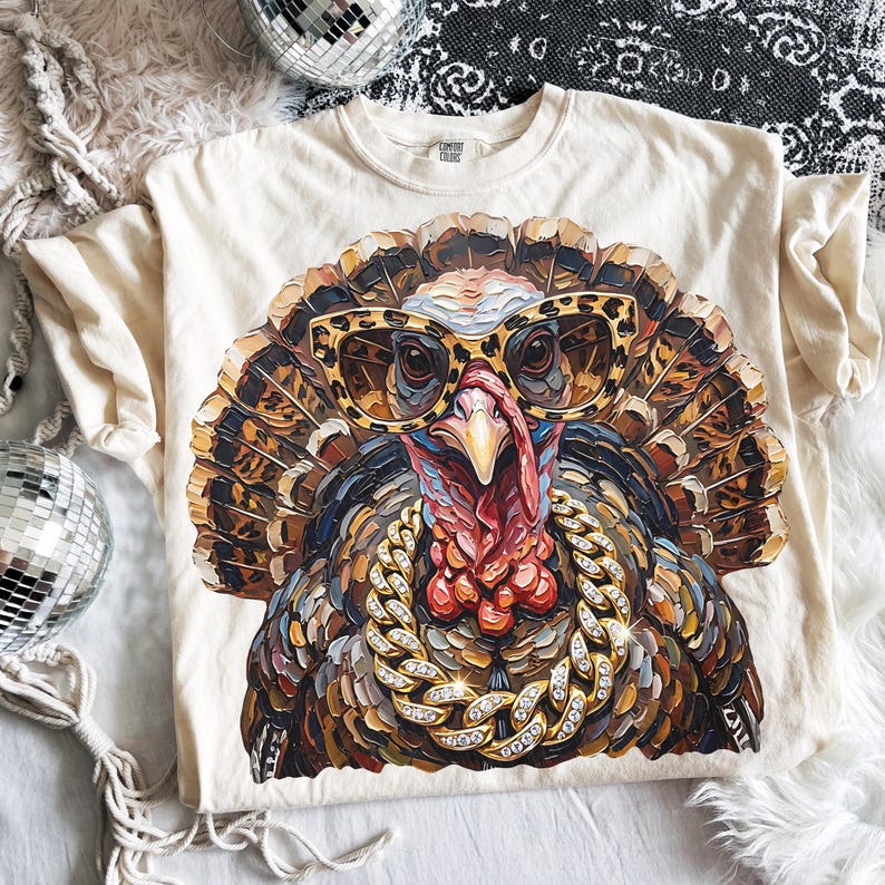 May include: A cream-colored t-shirt featuring a graphic print of a turkey wearing leopard-print sunglasses and a thick, gold-toned chain necklace with embedded crystals. The turkey is rendered in a textured, painterly style with brown, tan, and red tones.