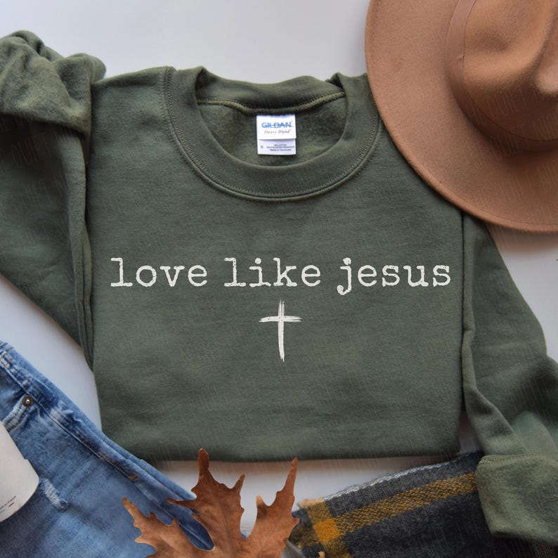 May include: An olive green sweatshirt with the text "love like jesus" in white, along with a white cross graphic. The sweatshirt is paired with blue jeans, a brown hat, and a plaid scarf.