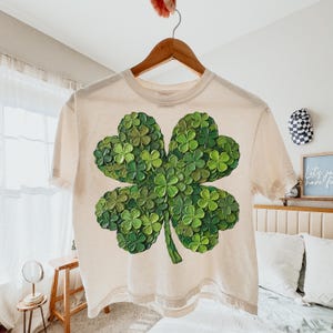 May include: Cream-colored t-shirt featuring a large, detailed four-leaf clover design. The clover is composed of numerous small green clovers, creating a textured, three-dimensional effect. The shirt is hanging on a wooden hanger.