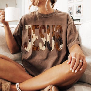 K&ouml;nnte beinhalten: Ein braunes T-Shirt mit dem Wort "JESUS" im Kuhfellmuster. Das Shirt hat eine lockere Passform und kurze &Auml;rmel. Die Person h&auml;lt eine wei&szlig;e Tasse.