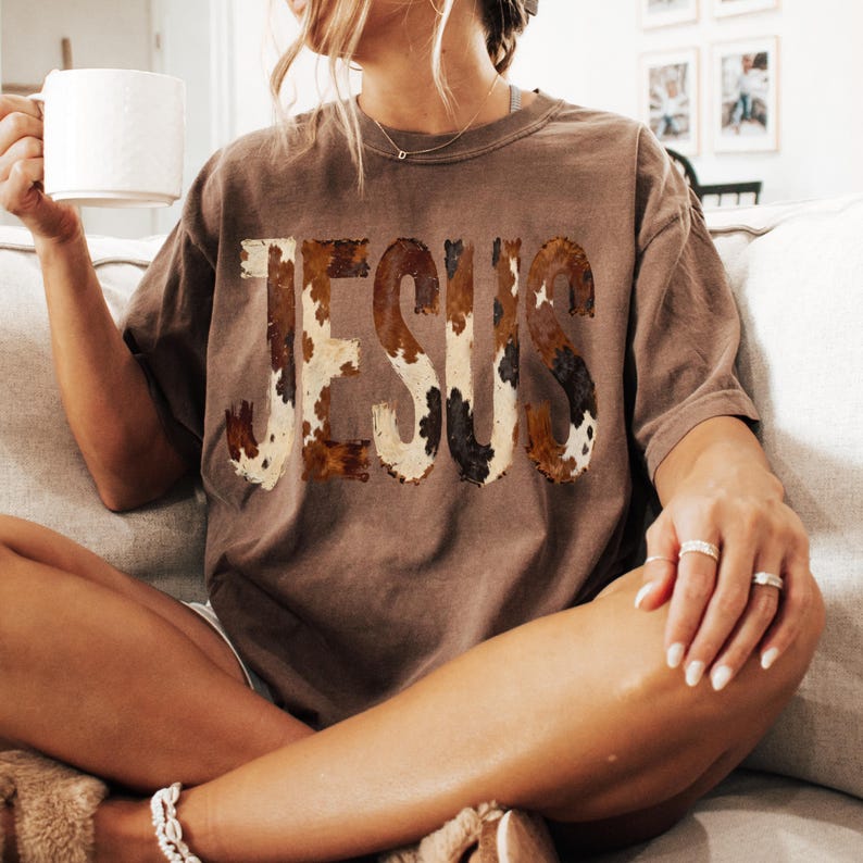 K&ouml;nnte beinhalten: Ein braunes T-Shirt mit dem Wort "JESUS" im Kuhfellmuster. Das Shirt hat eine lockere Passform und kurze &Auml;rmel. Die Person h&auml;lt eine wei&szlig;e Tasse.