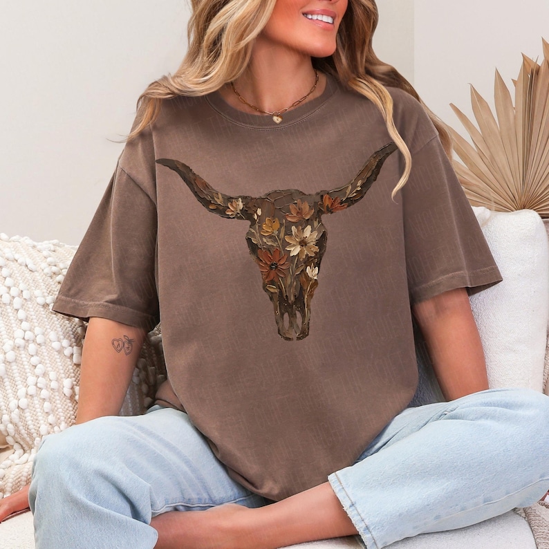 May include: Brown t-shirt featuring a detailed illustration of a bull skull adorned with flowers in shades of brown, orange, and cream. The shirt has short sleeves and a relaxed fit. The model is wearing light blue jeans.