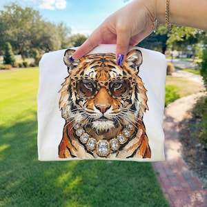 K&ouml;nnte beinhalten: Wei&szlig;es Sweatshirt mit einer detaillierten Illustration eines Tigers, der eine Brille mit Leopardenmuster und eine Diamantkette tr&auml;gt. Der Tiger hat orange und schwarze Streifen. Das Sweatshirt wird von einer Hand gehalten.