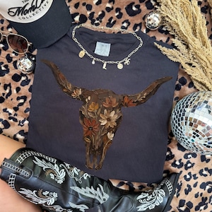 May include: A black t-shirt featuring a floral bull skull design. The image also includes a black baseball cap, sunglasses, a gold necklace with charms, a pair of black cowboy boots, and disco balls.