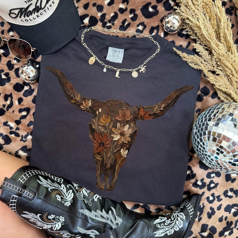 May include: A black t-shirt featuring a floral bull skull design. The image also includes a black baseball cap, sunglasses, a gold necklace with charms, a pair of black cowboy boots, and disco balls.