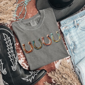 May include: A gray t-shirt with a horseshoe design in shades of brown, blue, and orange. The shirt is laid flat with a pair of blue jeans, a black cowboy boot, and a turquoise necklace. A black hat is also in the image.