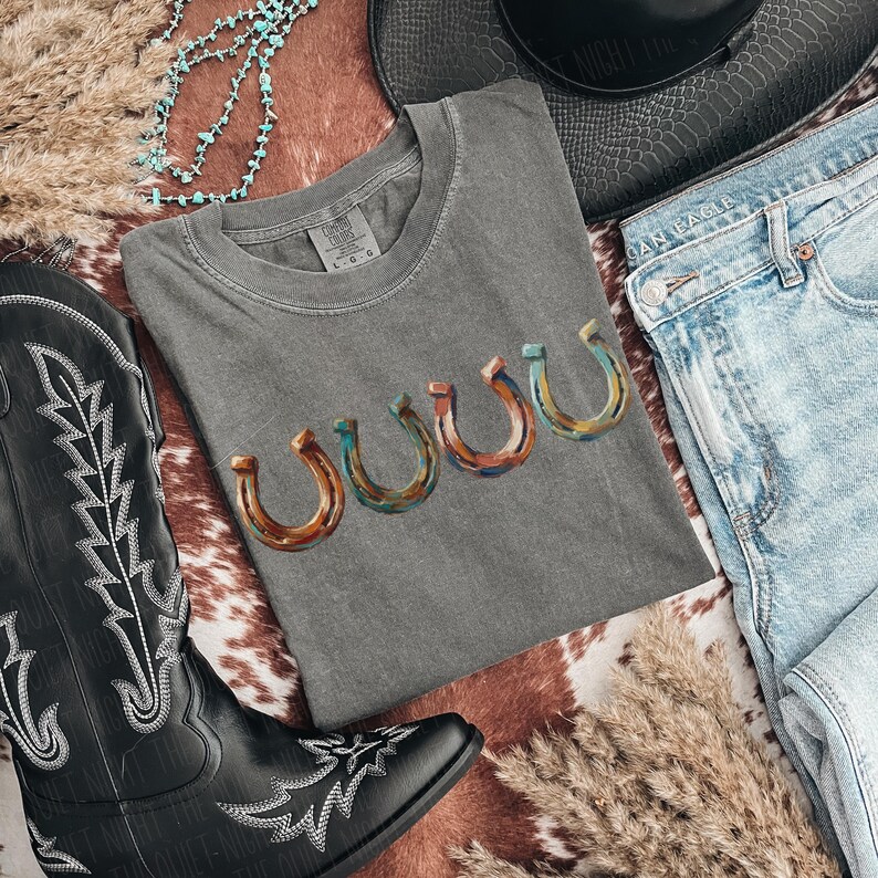 May include: A gray t-shirt with a horseshoe design in shades of brown, blue, and orange. The shirt is laid flat with a pair of blue jeans, a black cowboy boot, and a turquoise necklace. A black hat is also in the image.