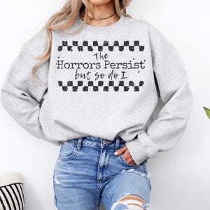 May include: A light gray sweatshirt with the text "The Horrors Persist but so do I" in black. The text is framed by a black and white checkered pattern. The sweatshirt is long-sleeved.