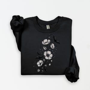 May include: Black sweatshirt featuring a floral design with white flowers and leaves. The design also includes several black bat silhouettes. The sweatshirt is a crew neck style.