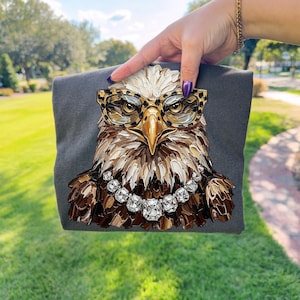 May include: A dark gray t-shirt featuring a detailed illustration of an eagle wearing leopard print glasses and a diamond necklace. The eagle's feathers are rendered in shades of brown, white, and gold.