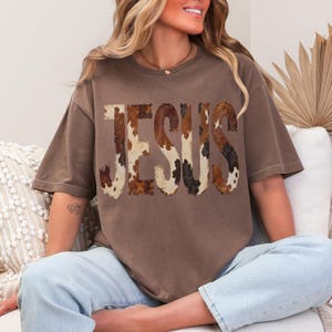 K&ouml;nnte beinhalten: Ein braunes T-Shirt mit dem Wort "JESUS" in einem Kuhfellmuster auf der Vorderseite. Das Shirt hat kurze &Auml;rmel und eine lockere Passform. Das Model tr&auml;gt hellblaue Jeans.