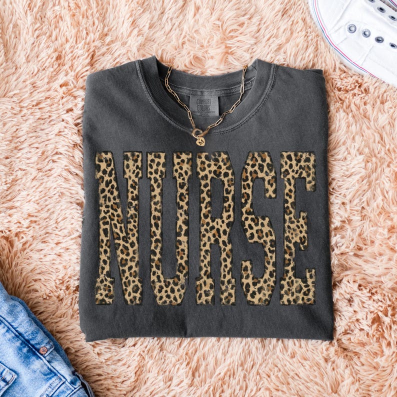 May include: Dark gray t-shirt with the word "NURSE" in large leopard print letters. A gold necklace and white sneakers are visible. The shirt is folded on a pink, fluffy surface.
