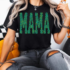K&ouml;nnte beinhalten: Ein schwarzes T-Shirt mit kurzen &Auml;rmeln und dem Wort MAMA in einer fetten Blockschrift. Die Buchstaben sind mit einem leuchtend gr&uuml;nen und gelben Leopardenmuster gef&uuml;llt, was ein kontrastreiches Grafikdesign auf der Vorderseite des Shirts ergibt.