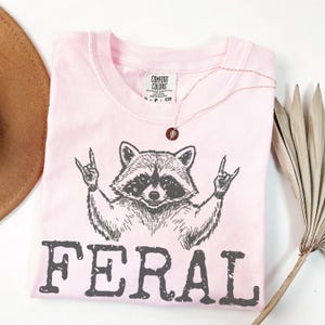 May include: A light pink t-shirt with a raccoon graphic giving a rock and roll hand gesture. The word "FERAL" is printed in dark gray below the raccoon. A gold necklace with a pendant is draped on the shirt.