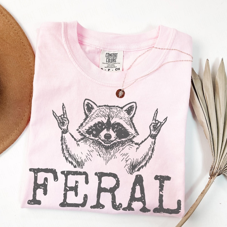 May include: A light pink t-shirt with a raccoon graphic giving a rock and roll hand gesture. The word "FERAL" is printed in dark gray below the raccoon. A gold necklace with a pendant is draped on the shirt.