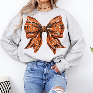 May include: A light gray sweatshirt featuring a large bow design. The bow is orange and black, with a basketball court pattern. The sweatshirt is a crew neck style and is made of a soft material.