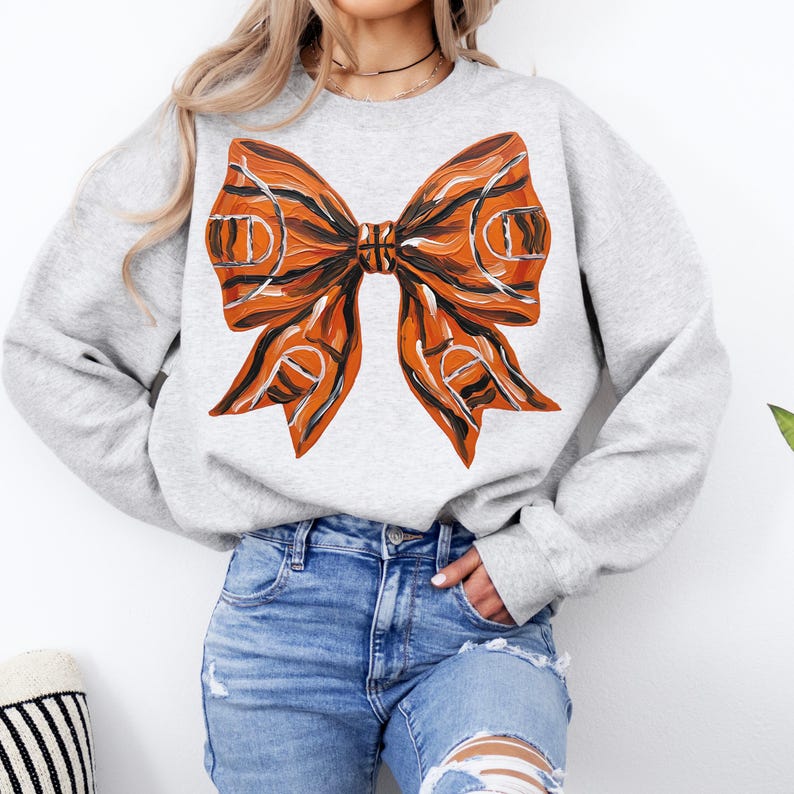 May include: A light gray sweatshirt featuring a large bow design. The bow is orange and black, with a basketball court pattern. The sweatshirt is a crew neck style and is made of a soft material.