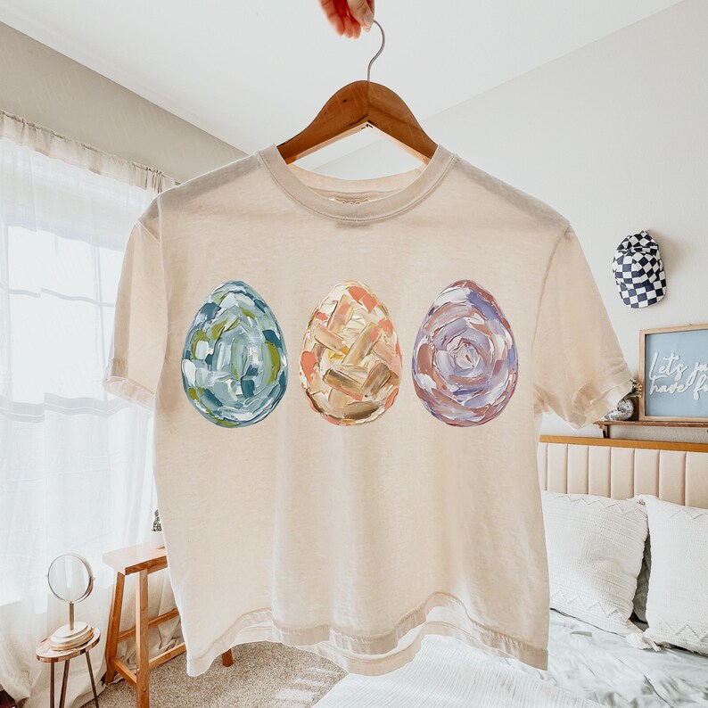 Peut inclure: Un t-shirt cr&egrave;me avec trois &oelig;ufs de P&acirc;ques peints. Les &oelig;ufs sont dans des tons de bleu, d'orange et de violet, avec un motif textur&eacute; au pinceau. Le t-shirt est suspendu &agrave; un cintre en bois.