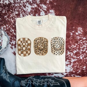 以下が含まれることがあります： 3つのカウボーイハットのデザインが施されたクリーム色のTシャツ。ハットは市松模様、ヒョウ柄、水玉模様です。Tシャツは赤と茶色のカウハイド柄の上に折りたたまれています。