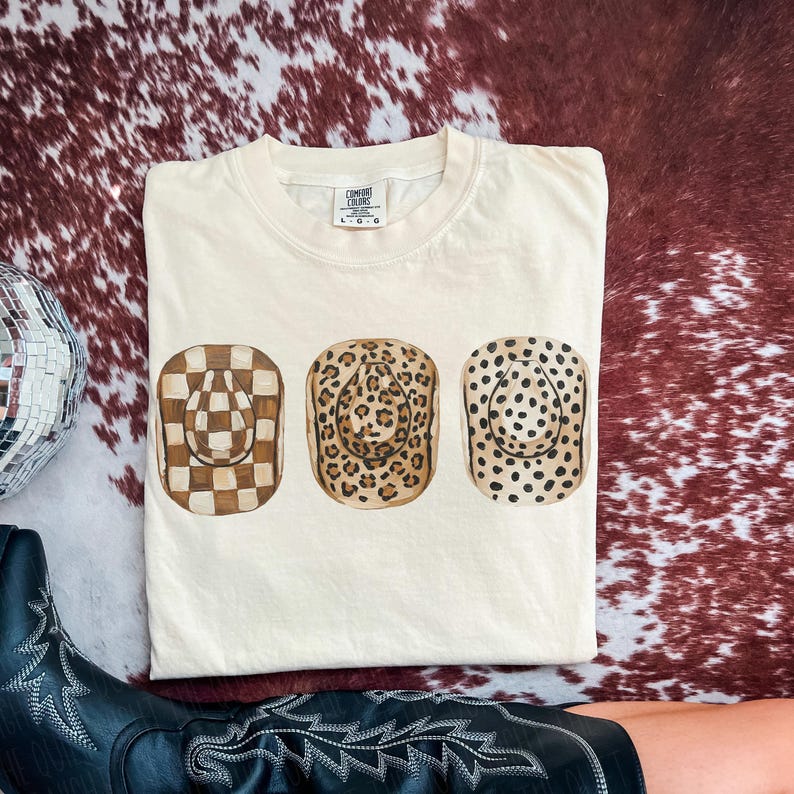 以下が含まれることがあります： 3つのカウボーイハットのデザインが施されたクリーム色のTシャツ。ハットは市松模様、ヒョウ柄、水玉模様です。Tシャツは赤と茶色のカウハイド柄の上に折りたたまれています。
