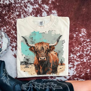 May include: Cream-coloured t-shirt with a detailed illustration of a Highland cow with long horns. The graphic includes a desert landscape with cacti and a turquoise and grey background. The shirt is labelled "Comfort Colors".