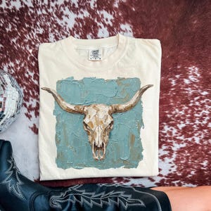 May include: Cream Comfort Colors t-shirt with a bull skull design on a textured teal background. The bull skull is painted in brown and cream shades. The shirt is folded on a cowhide patterned surface.