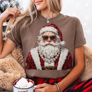 May include: A brown short-sleeve t-shirt featuring a graphic print of Santa Claus wearing leopard print sunglasses and a thick gold chain. The Santa illustration has a textured, painterly style with a red hat, white beard, and red coat.