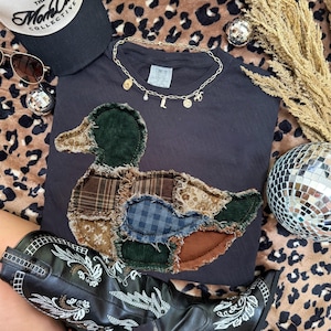 May include: A dark gray t-shirt featuring a large, central duck-shaped appliqu&eacute; made from various fabric patches, including green corduroy, brown plaid, blue gingham, and floral prints with frayed edges. A gold-toned charm necklace rests above.