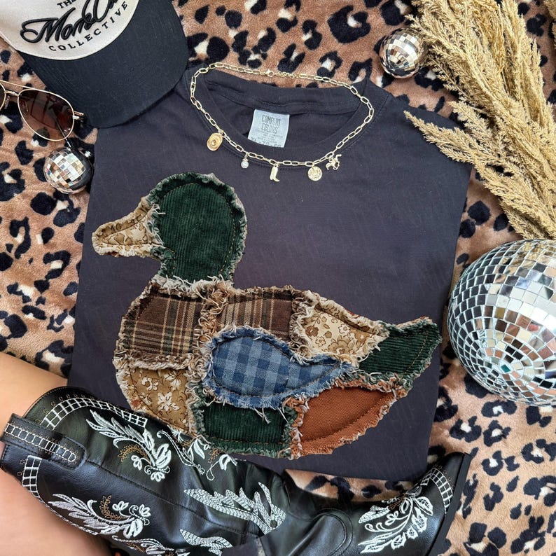 May include: A dark gray t-shirt featuring a large, central duck-shaped appliqu&eacute; made from various fabric patches, including green corduroy, brown plaid, blue gingham, and floral prints with frayed edges. A gold-toned charm necklace rests above.