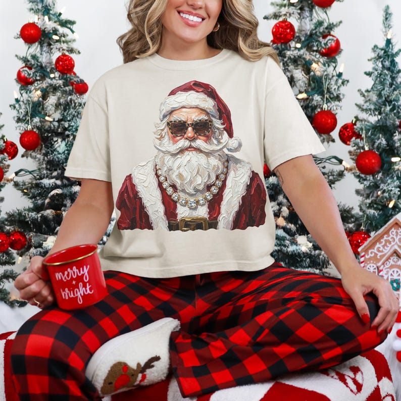 May include: A cream-colored t-shirt featuring a graphic of Santa Claus wearing leopard-print sunglasses and a rhinestone necklace. The Santa illustration is rendered in a textured, painterly style. A red mug with the text merry bright is held.