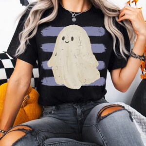 May include: A black short-sleeve t-shirt featuring a central graphic of a cream-colored ghost with a simple smiling face. The ghost is positioned over four horizontal, distressed purple brushstroke stripes that span the width of the chest area.