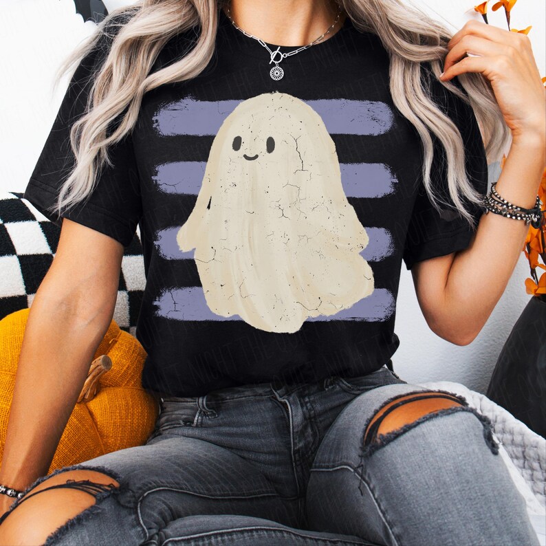 May include: A black short-sleeve t-shirt featuring a central graphic of a cream-colored ghost with a simple smiling face. The ghost is positioned over four horizontal, distressed purple brushstroke stripes that span the width of the chest area.