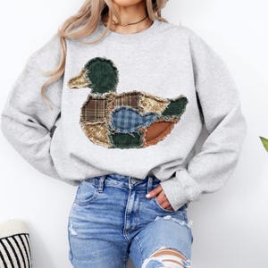 May include: A light gray crewneck sweatshirt featuring a large central applique of a duck. The duck is constructed from various fabric patches including green corduroy, brown plaid, blue gingham, and floral patterns with frayed edges.