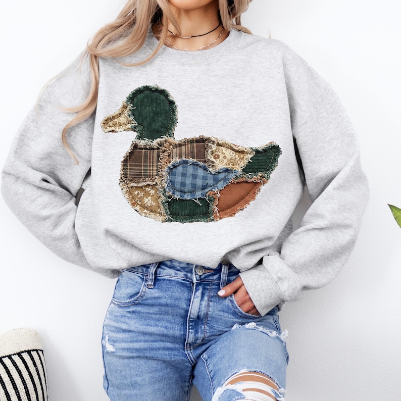 May include: A light gray crewneck sweatshirt featuring a large central applique of a duck. The duck is constructed from various fabric patches including green corduroy, brown plaid, blue gingham, and floral patterns with frayed edges.