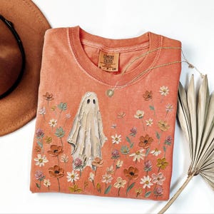 Może przedstawiać: T-shirt z okrągłym dekoltem w kolorze terakoty z centralną grafiką przedstawiającą białego ducha otoczonego polem malowanych kwiat&oacute;w w odcieniach r&oacute;żu, bieli i pomarańczu. Koszulka posiada metkę marki Comfort Colors przy wewnętrznym dekolcie.