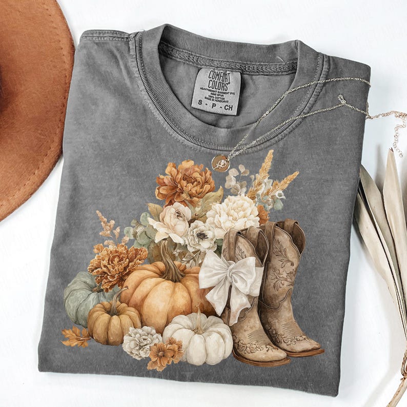 May include: Gray Comfort Colors t-shirt featuring a fall-themed graphic. The design includes pumpkins, flowers, and cowboy boots. A silver necklace with a pendant is draped across the shirt. The shirt is a size small.