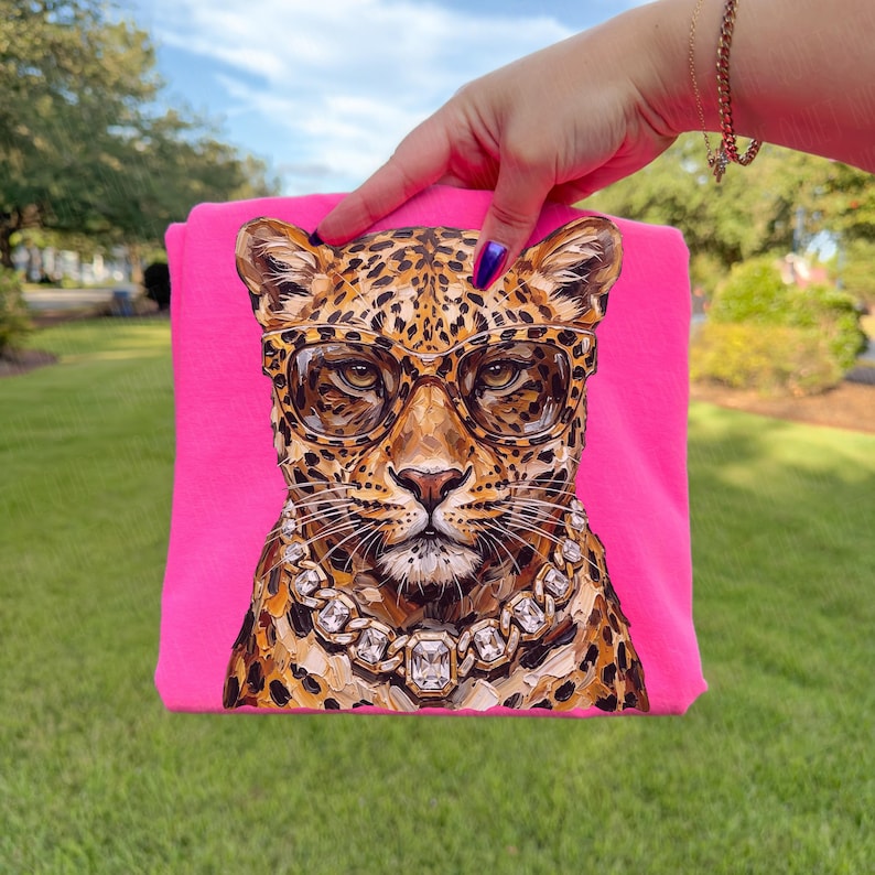 Puede incluir: Una camiseta rosa intenso con una ilustraci&oacute;n detallada de un leopardo con gafas y un collar de diamantes. El leopardo tiene un pelaje manchado y est&aacute; centrado en la camiseta. La camiseta est&aacute; sujeta por una persona.