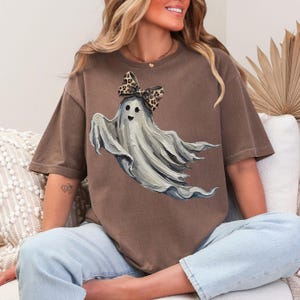 May include: Brown t-shirt with a white ghost illustration wearing a leopard print bow. The shirt has a loose fit, ideal for Halloween or everyday wear. The design is printed on a comfortable fabric.